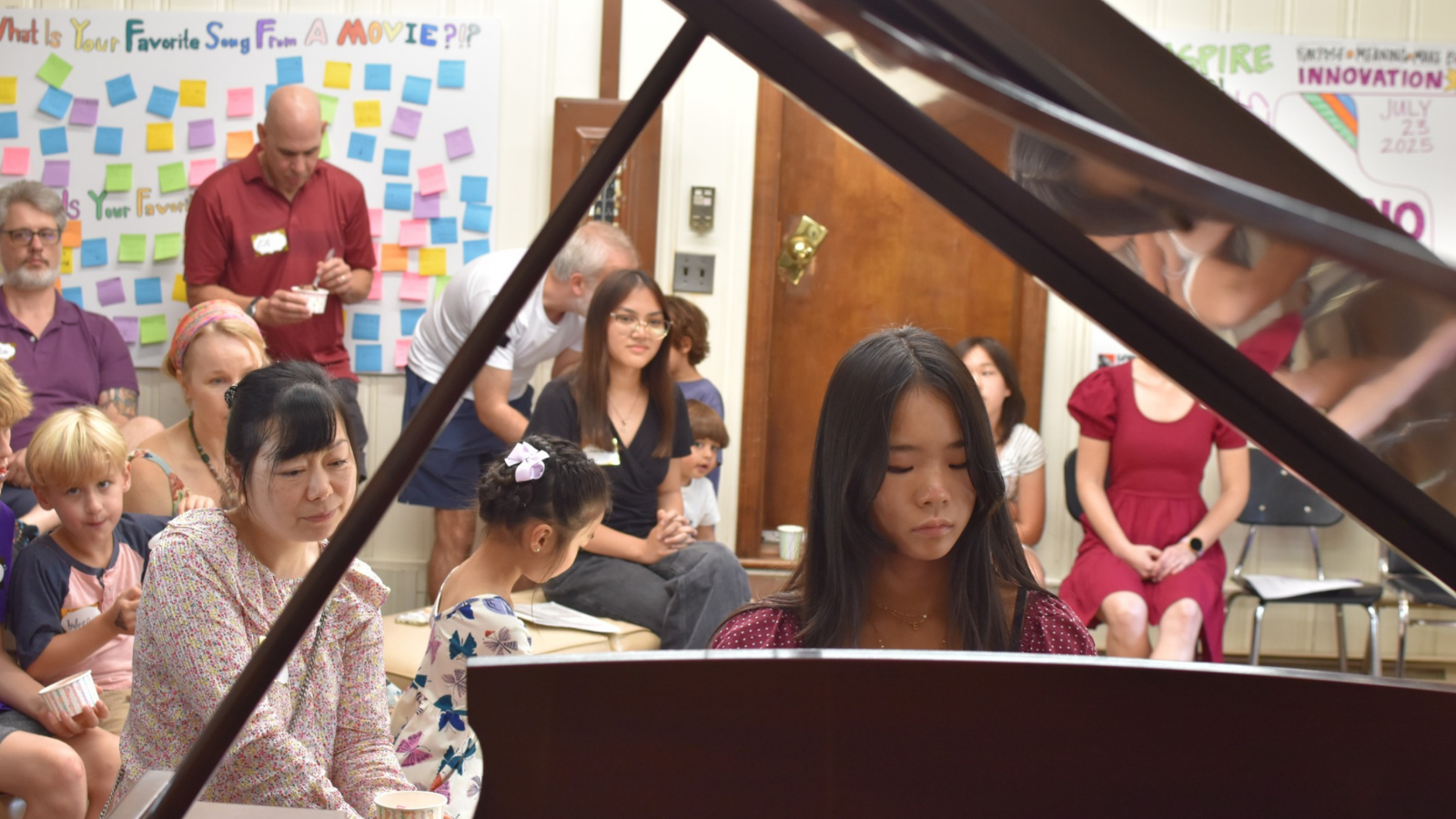 Woman playing a piano in a classroom with children and adults watching.