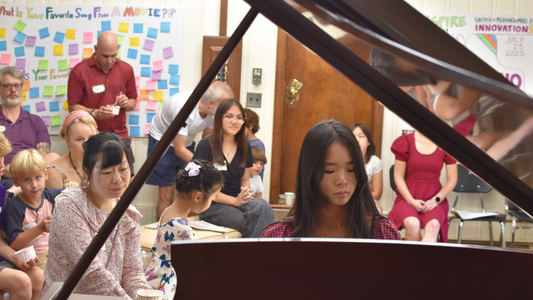 Woman playing a piano in a classroom with children and adults watching.