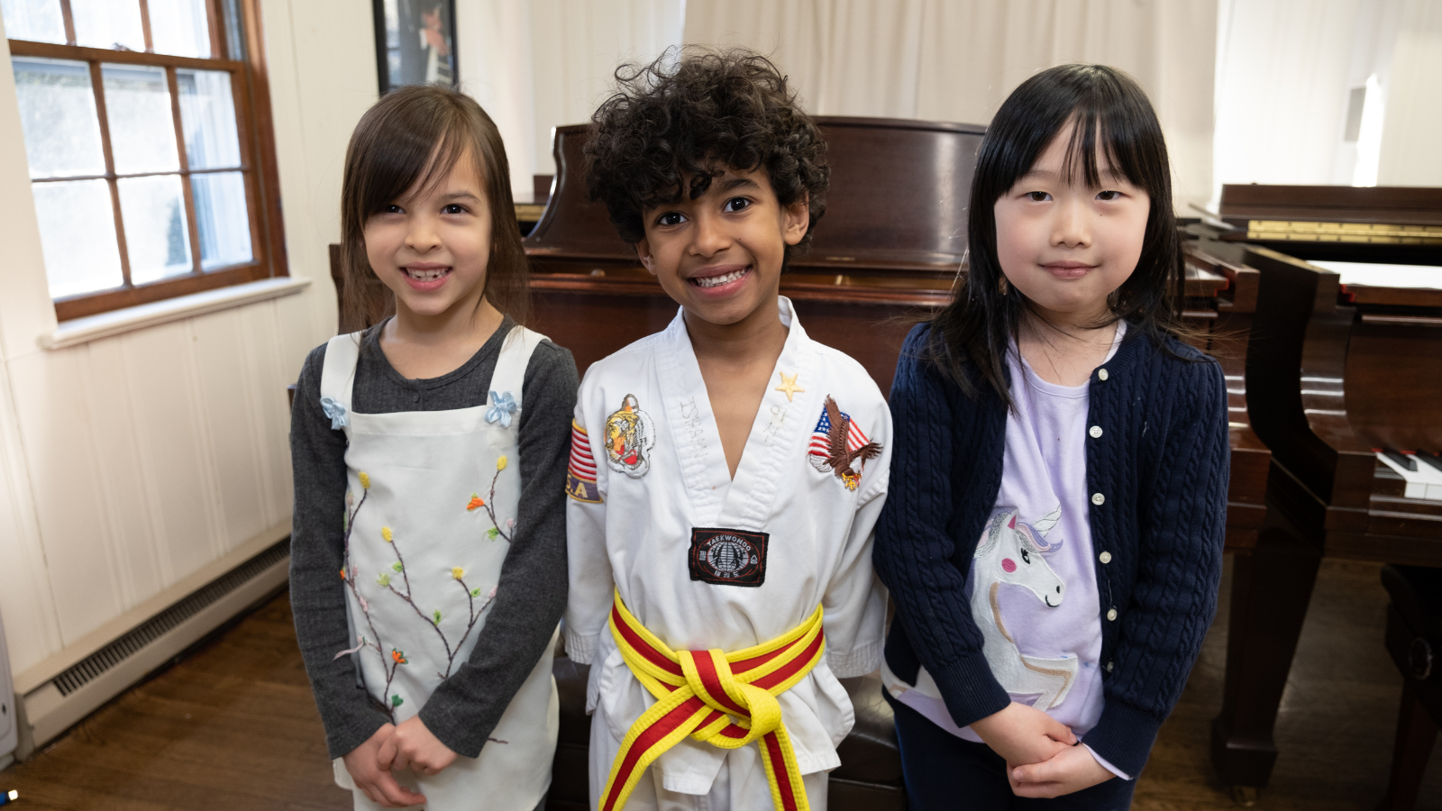 Three children standing in a room with a piano, one wearing a martial arts uniform with a yellow belt.