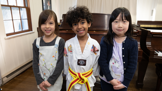 Three children standing in a room with a piano, one wearing a martial arts uniform with a yellow belt.